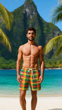 Man wearing colorful swim shorts standing on a tropical beach with mountains and palm trees in the background.