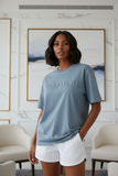 Woman wearing a blue t-shirt with embossed "pas jalou" text in a room with white walls and beige chairs.