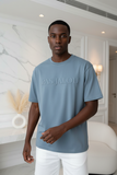 Man wearing a blue t-shirt with 'PAS JALOU' text in a modern indoor setting