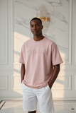 Man wearing a pink peachy  t-shirt with "pas jalou" embossed text and white shorts in a well-lit room.
