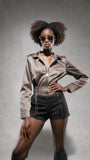 Woman wearing PAS JALOU grey sunglasses, a beige jacket and black shorts against a gray background
