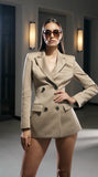 Woman in a beige suit and pas jalou sunglasses standing in a dimly lit room.