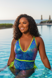 Woman in a colorful sequin swimsuit holding a glass of champagne by the water at sunset.