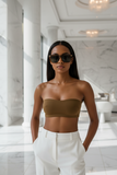 Woman wearing pas jalou sunglasses and also  a brown strapless top and white pants in a marble interior setting. 