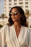 Woman wearing Pas Jalou sunglasses and a white outfit with a cityscape and palm trees in the background