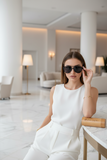 Woman in a white outfit sitting in a modern, well-lit room, wearing pas Jalou sunglasses