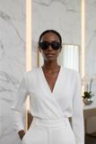 Woman in a white outfit wearing sunglasses, standing in a room with marble walls and a desk.