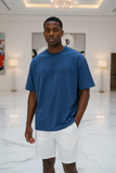 Man wearing a blue t-shirt with 'PAS JALOU' branding in a white room.