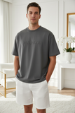 Man wearing a gray t-shirt with 'PAS JALOU' logo and white shorts in a bright room.