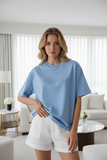 Woman wearing a blue oversized t-shirt with 'PAS JALOU' text in a modern living room.