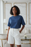 Woman wearing a blue t-shirt with embossed "pas jalou" text and white shorts in an elegant room.