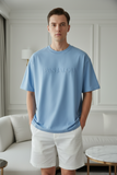 Man wearing a blue t-shirt with 'PAS JALOU' branding in a living room.
