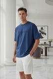 Man wearing a blue t-shirt with 'PAS JALOU' logo in a modern indoor setting